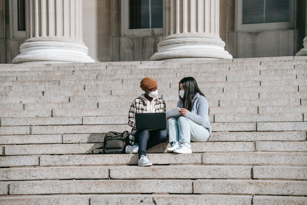 Two students sit on the steps of a building talking. One is holding a laptop.